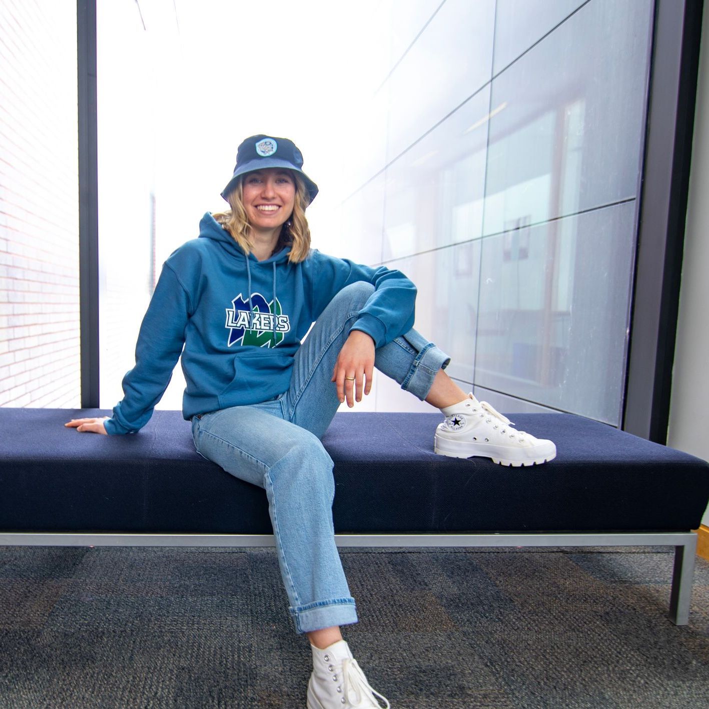 Woman in blue hoodie, jeans, and bucket hat smiles, sitting on a blue bench.