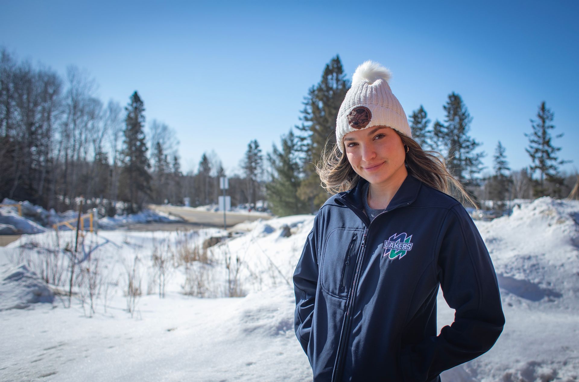 Woman in a beanie and jacket stands in a snowy, outdoor setting, smiling at the camera.