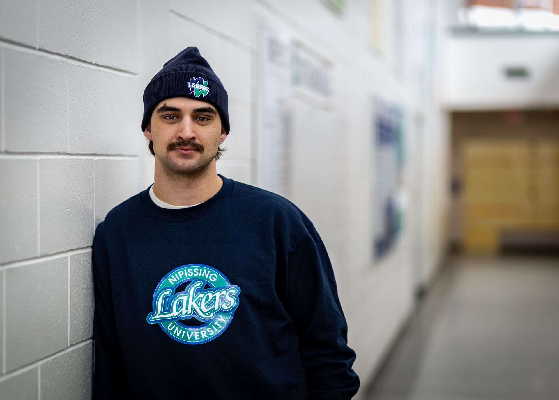 Man wearing a Lakers sweatshirt and beanie leaning against a wall in a hallway.