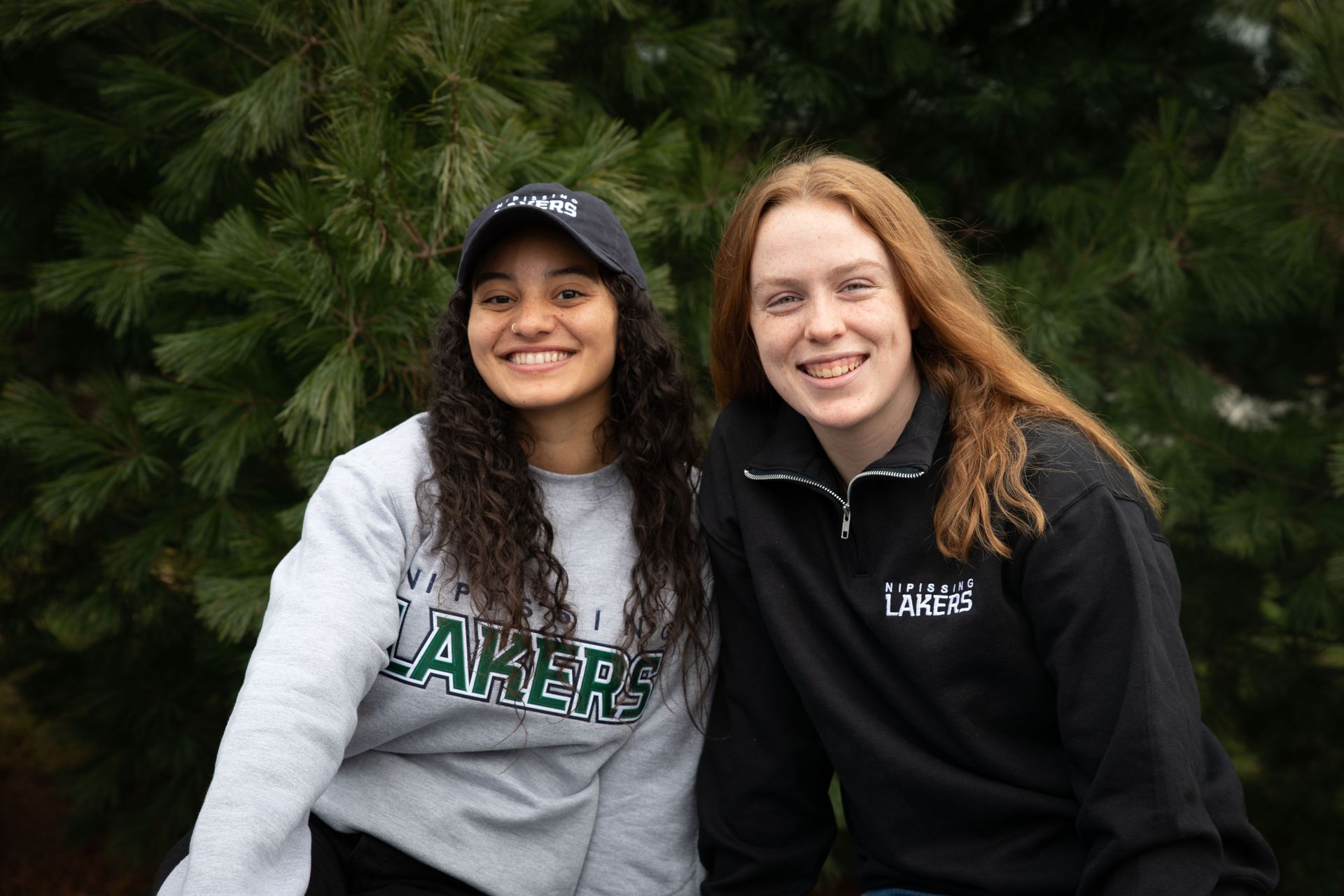 Two young women smiling in front of evergreen trees. One wears a black cap and sweatshirt, the other a grey sweatshirt.