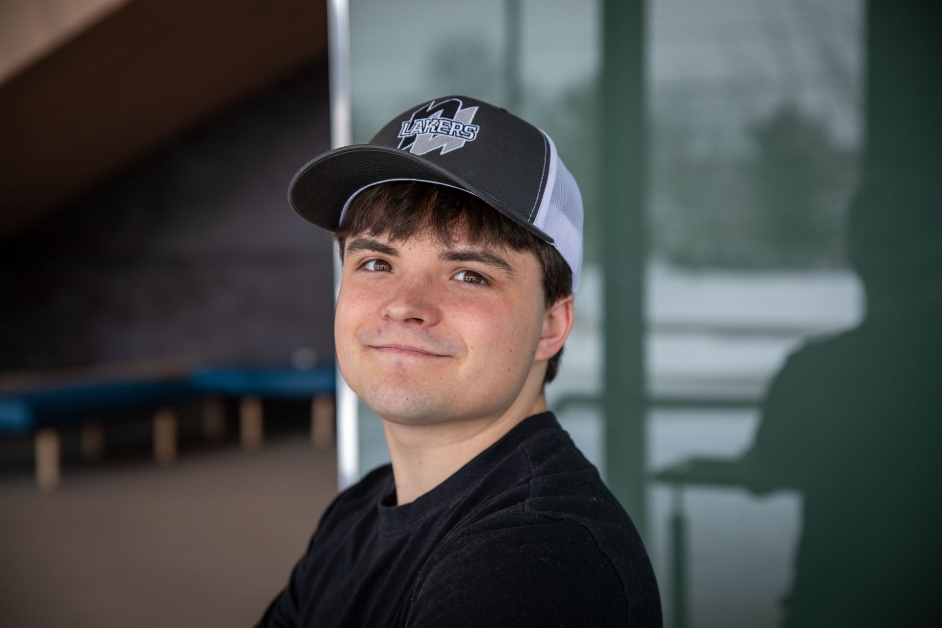 Young man with a baseball cap smiles at the camera. He wears a black shirt, standing indoors with glass panels.