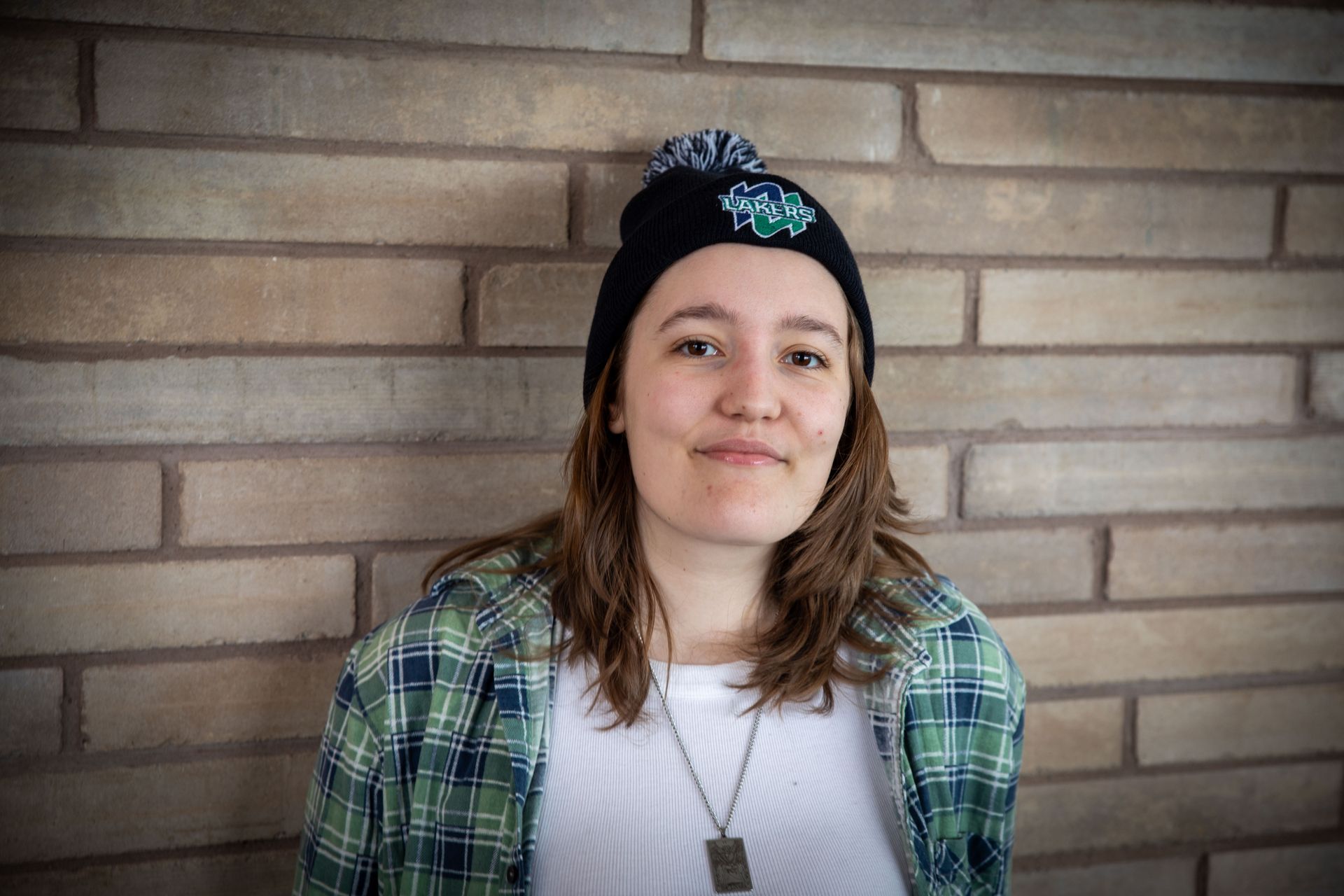Woman in beanie and flannel against a brick wall, smiling.