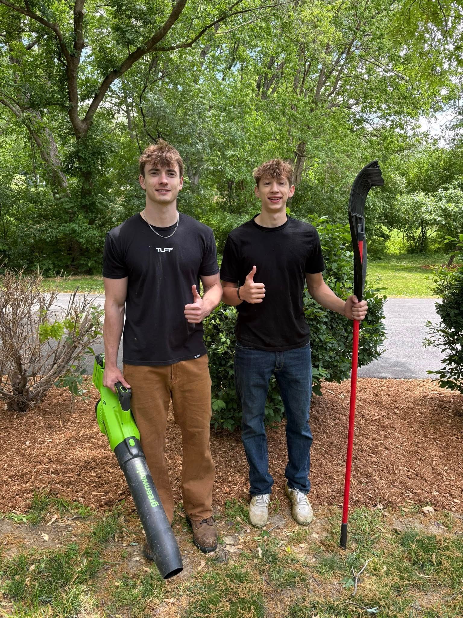 Two young men are standing next to each other holding lawn tools and giving a thumbs up.