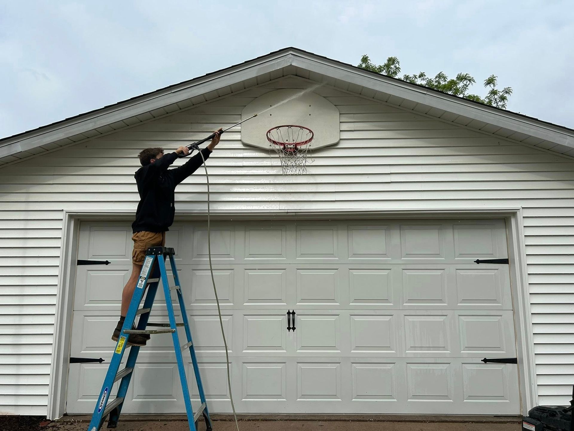 A man on a ladder is cleaning a basketball hoop on the side of a garage.