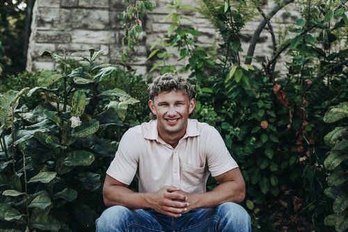 A man is sitting on the ground in front of a brick wall surrounded by plants.