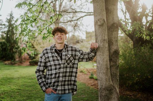 A young man in a plaid shirt is leaning against a tree.