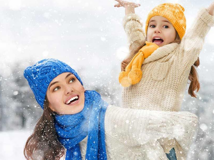 Woman and child play in the snow, both wearing knit hats and scarves, joyful expressions.