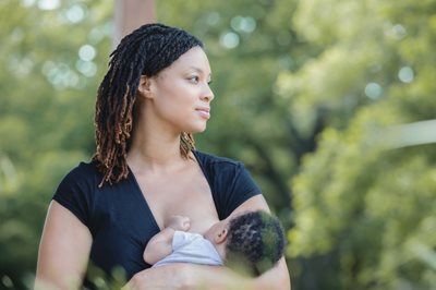 Woman breastfeeding outdoors, looking off-camera. Baby is held close. Green foliage in background.