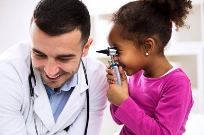 Doctor smiles while girl looks into an otoscope; doctor's office setting.