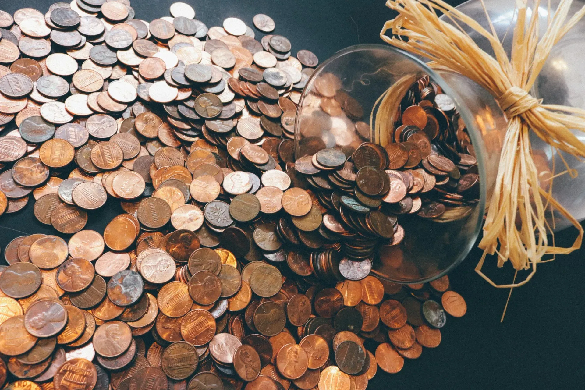 Pennies spilling from a glass jar, scattered on a dark surface.