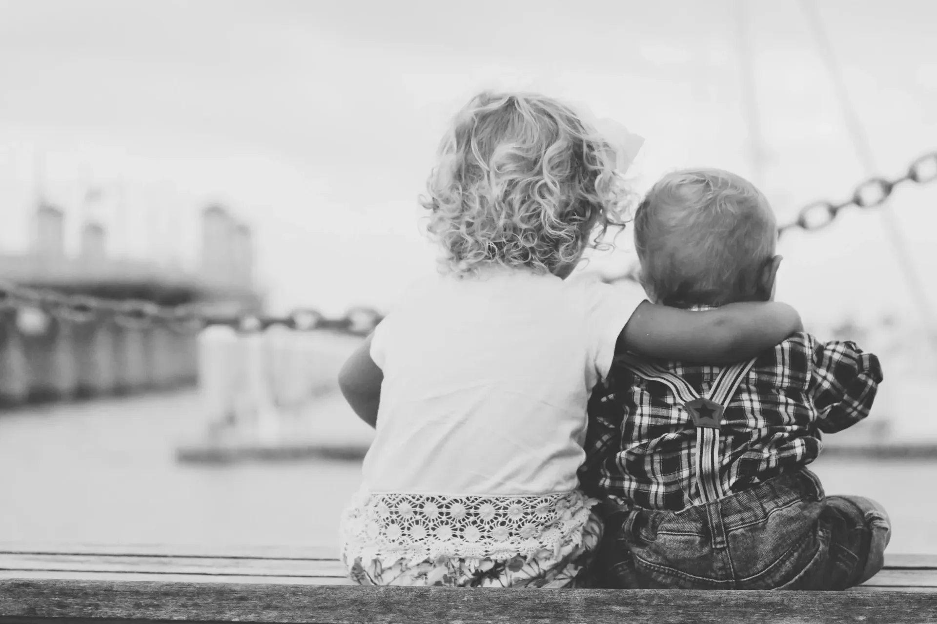Two young children sit side-by-side, hugging, looking out at a harbor.