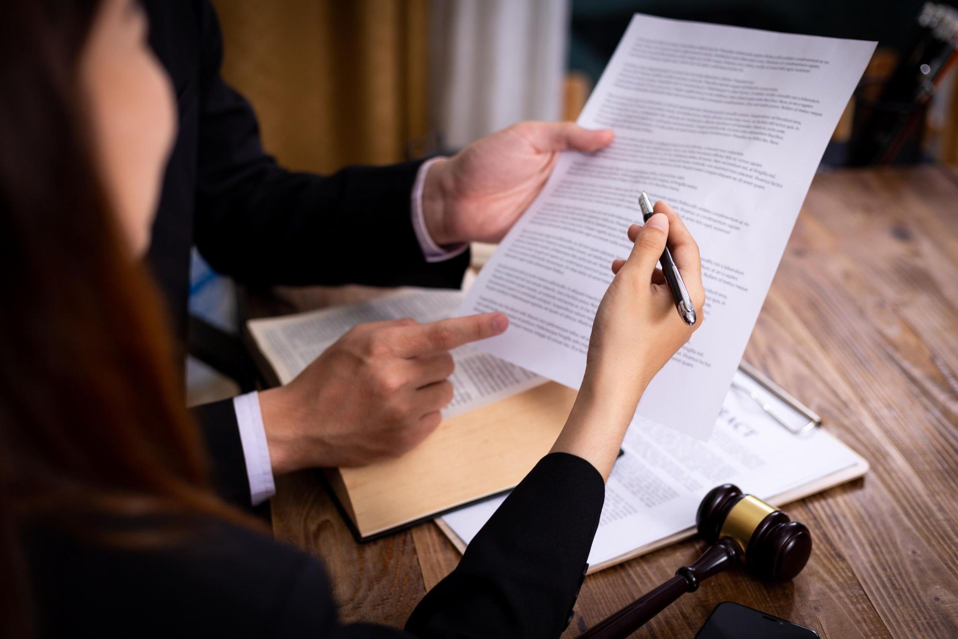 Two people in suits reviewing a document, one pointing, the other holding a pen; a gavel and open book on the desk.