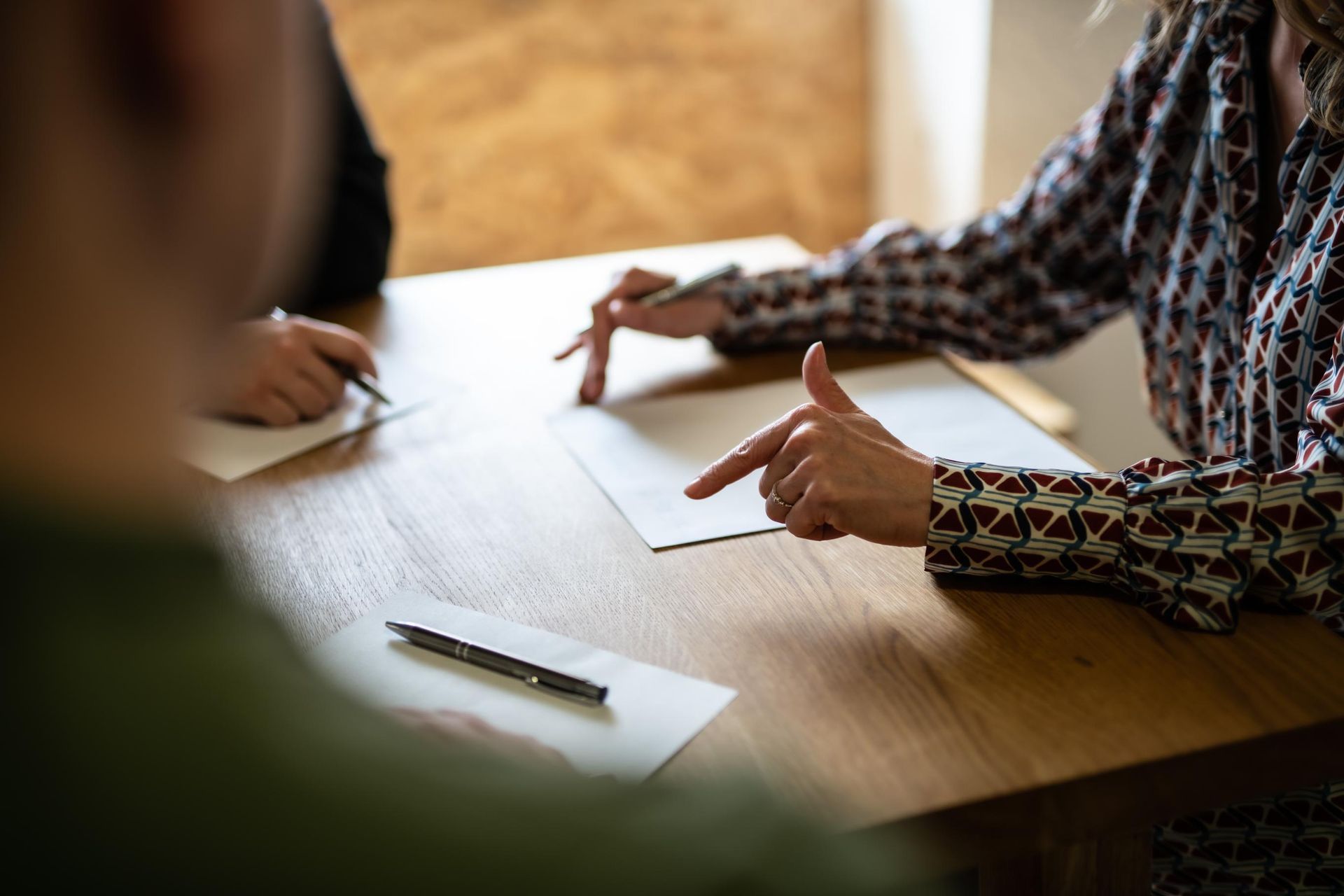 People at a table, pointing at blank papers. One writes with a pen. Wooden table, neutral tones.