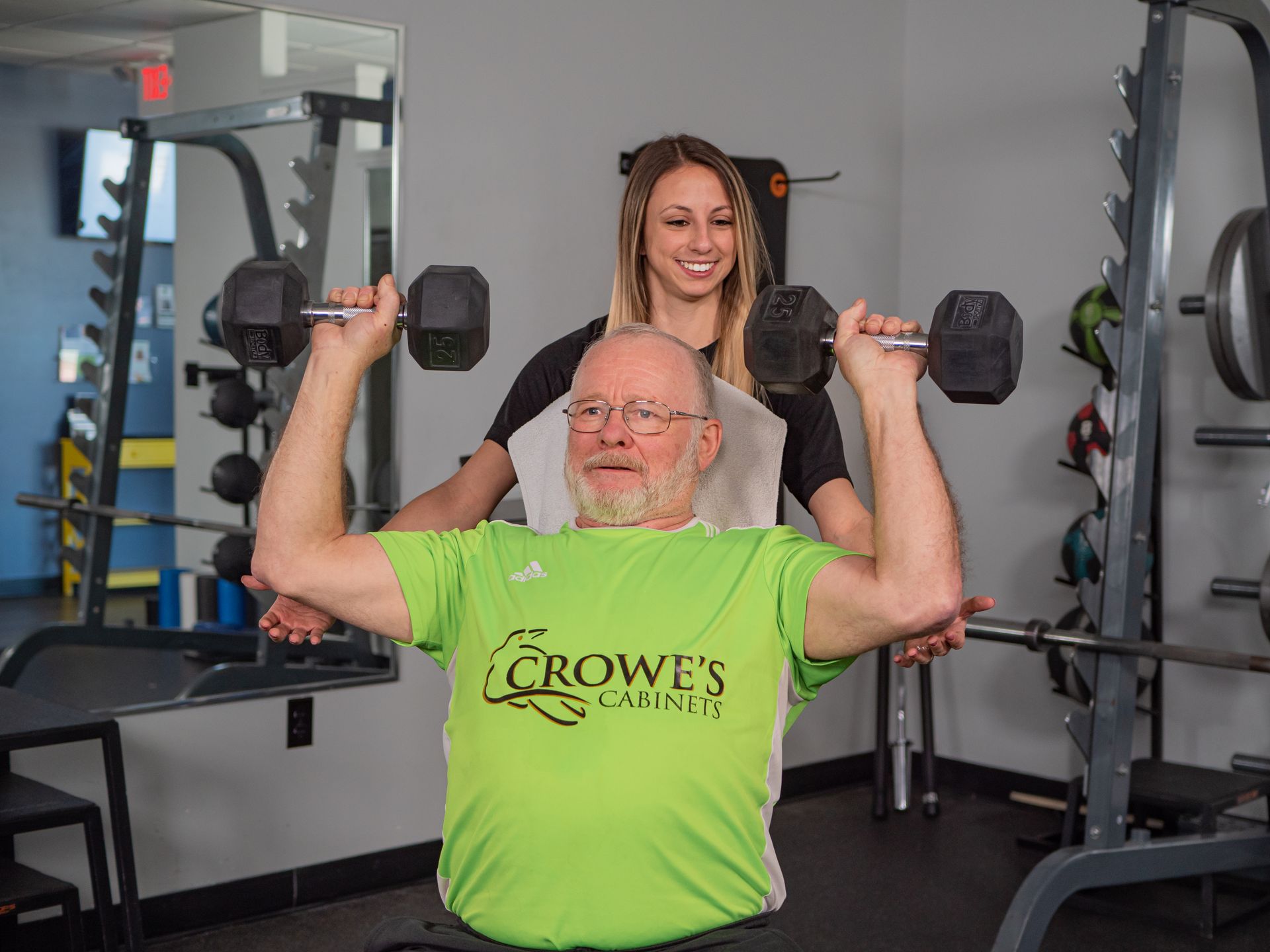 A personal trainer spots an older man lifting dumbbells overhead in a gym.
