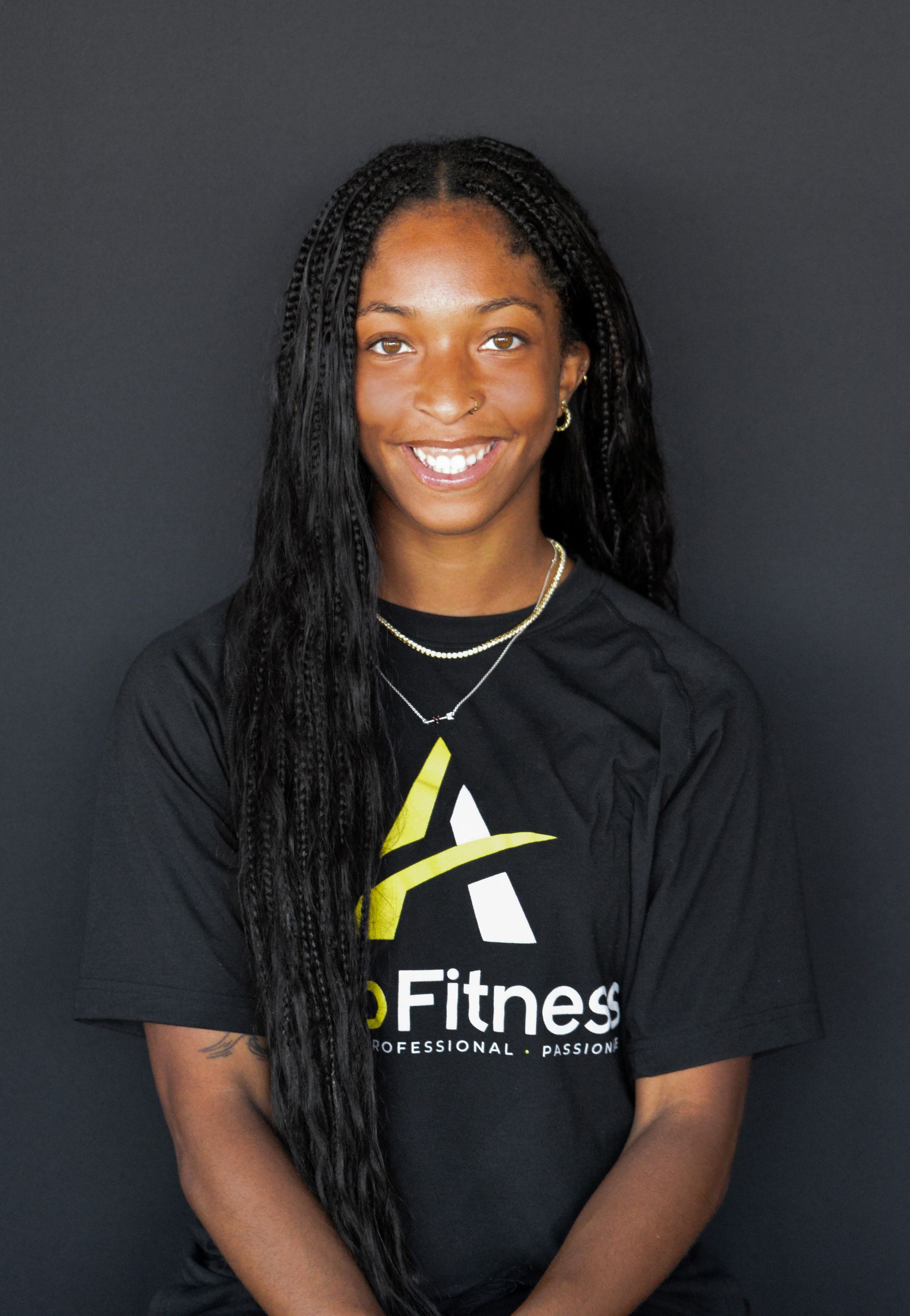 A young Black woman with long braids smiles. She wears a black t-shirt with a logo and jewelry, against a dark background.