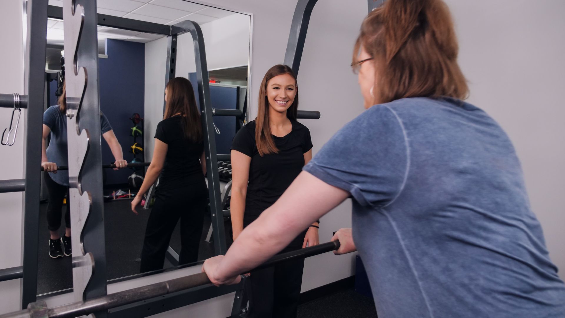 A woman in a gym uses a barbell with a trainer watching, smiling. The setting is a well-lit gym with a mirror.
