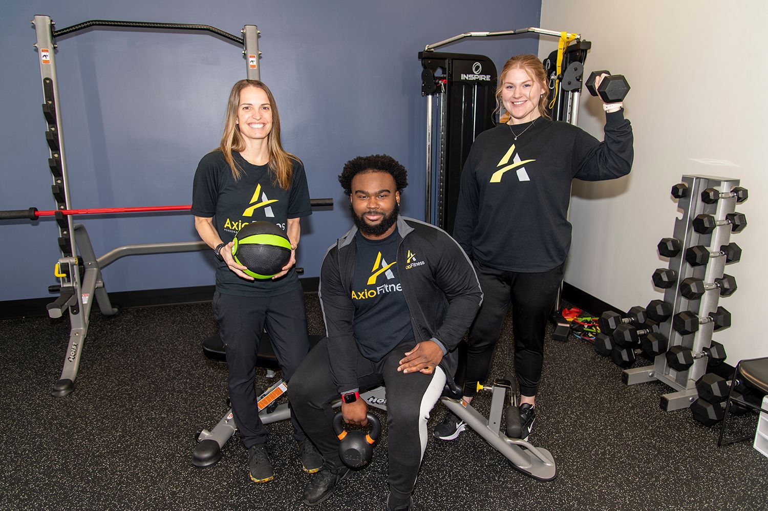 Three people in a gym, posing with fitness equipment. Two women and a man in black athletic wear.