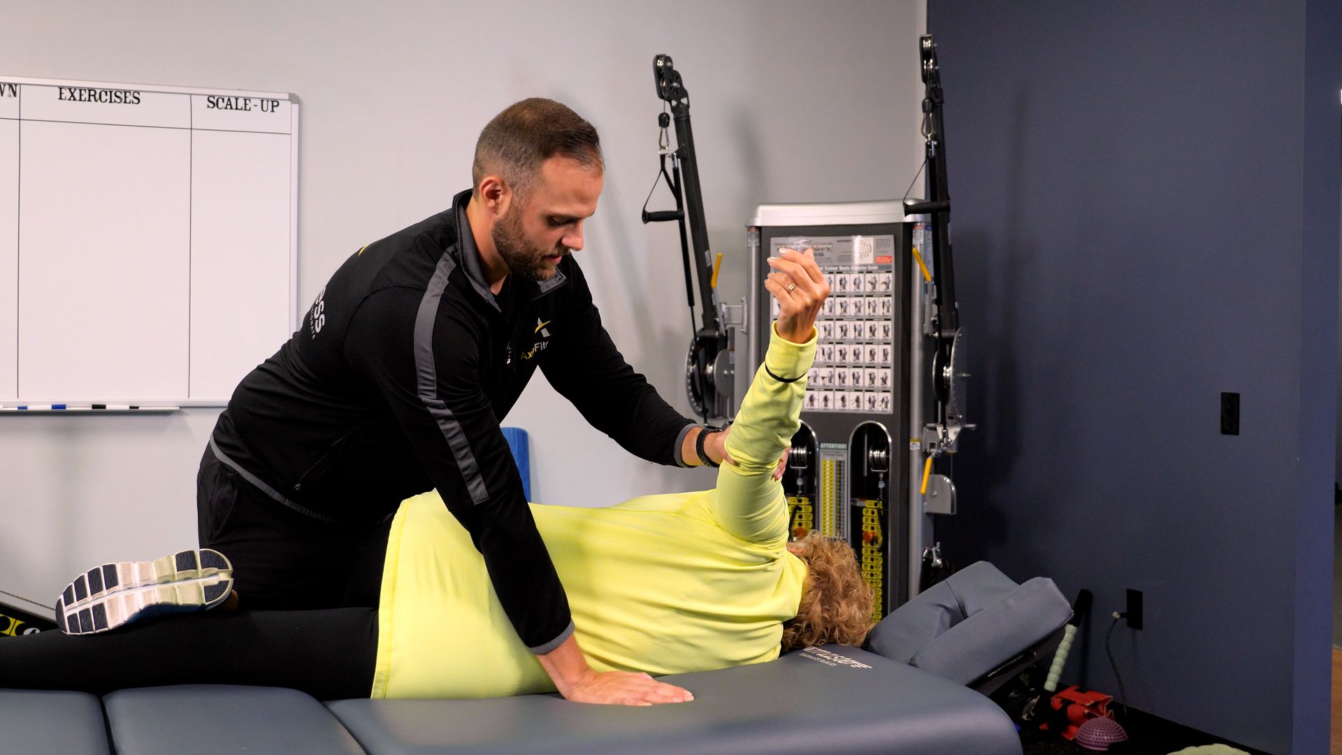 A man assists a woman doing an arm exercise while lying on a medical table. They are indoors.