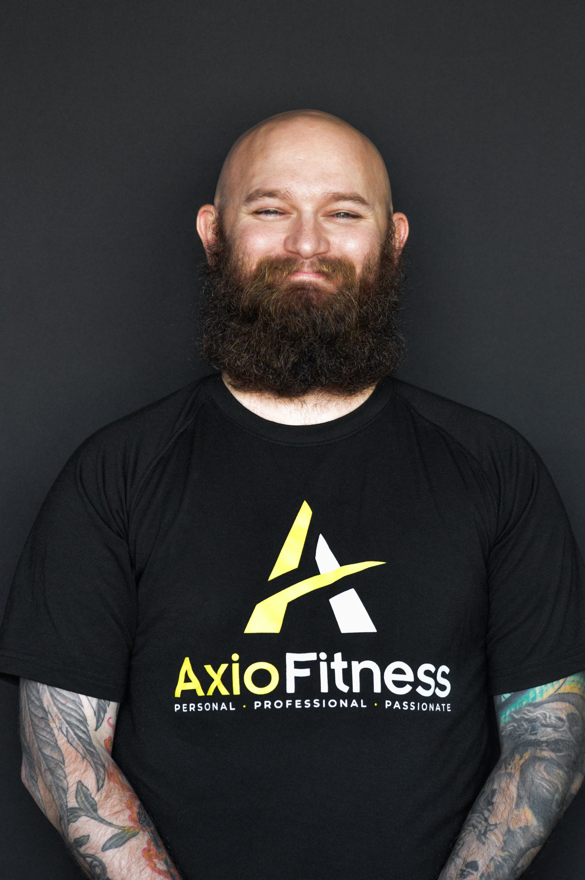 Man with a large beard, wearing a black AxioFitness t-shirt, smiling, against a gray background.