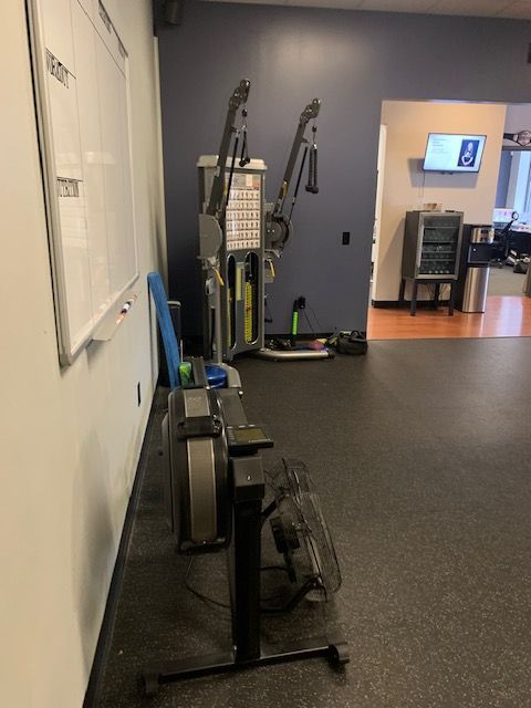 Rowing machine and weight machine in a gym. Blue wall, dark floor. Whiteboard on left.