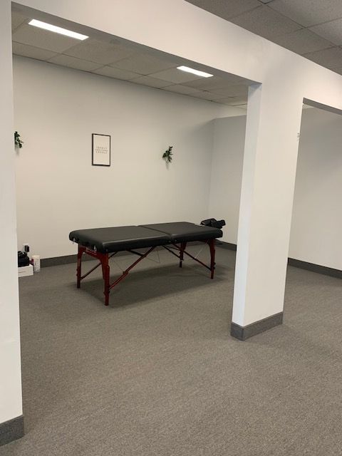 A massage therapy room with a black massage table, white walls, gray carpet, and a framed image.