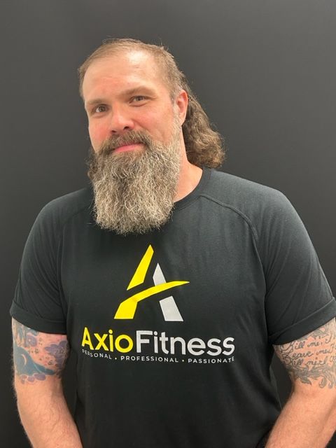 Man with a long beard wearing a black AxioFitness shirt, looking at the camera, with tattoos on his arms.