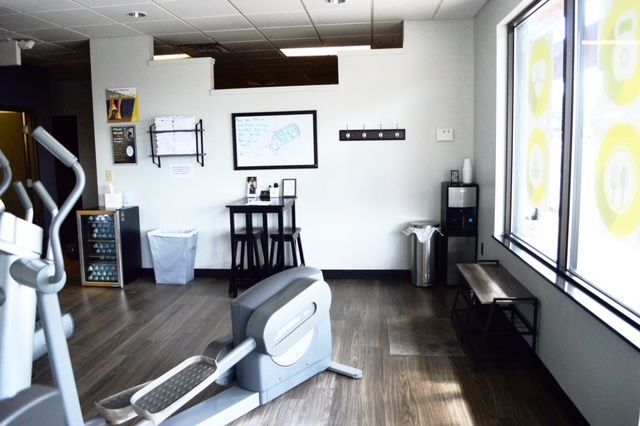 Elliptical machine in a gym with wooden floors, a small table, a water cooler, and a window with signage.