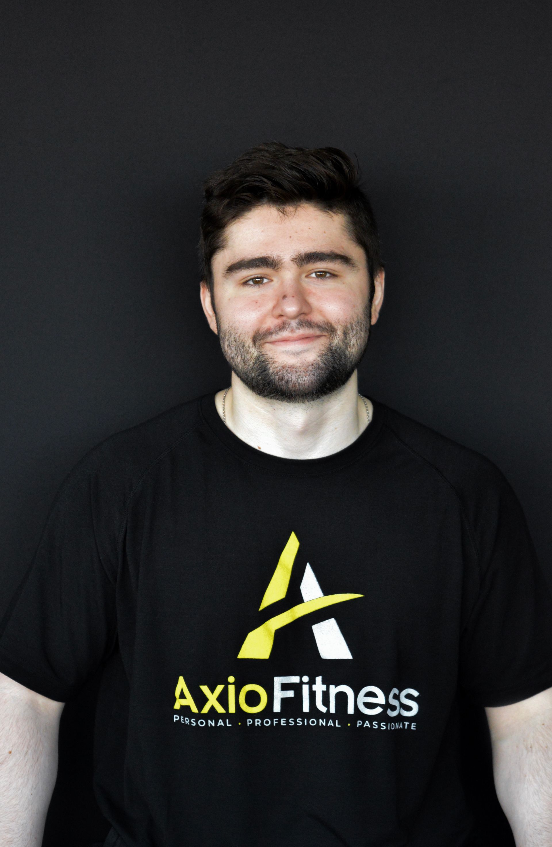 Man with dark hair and beard wearing a black AxioFitness t-shirt, smiling in front of a black backdrop.