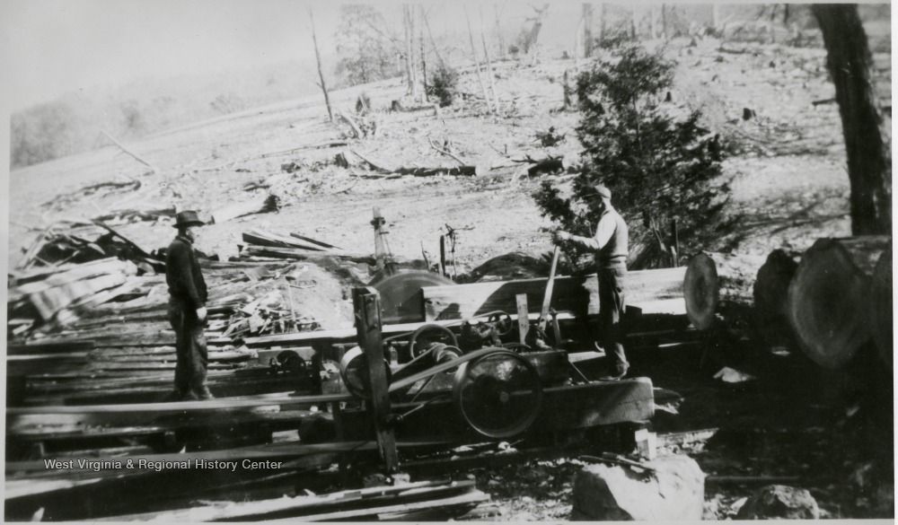 Two people operate a circular sawmill, cutting logs into lumber in a clearing with scattered trees and felled logs.