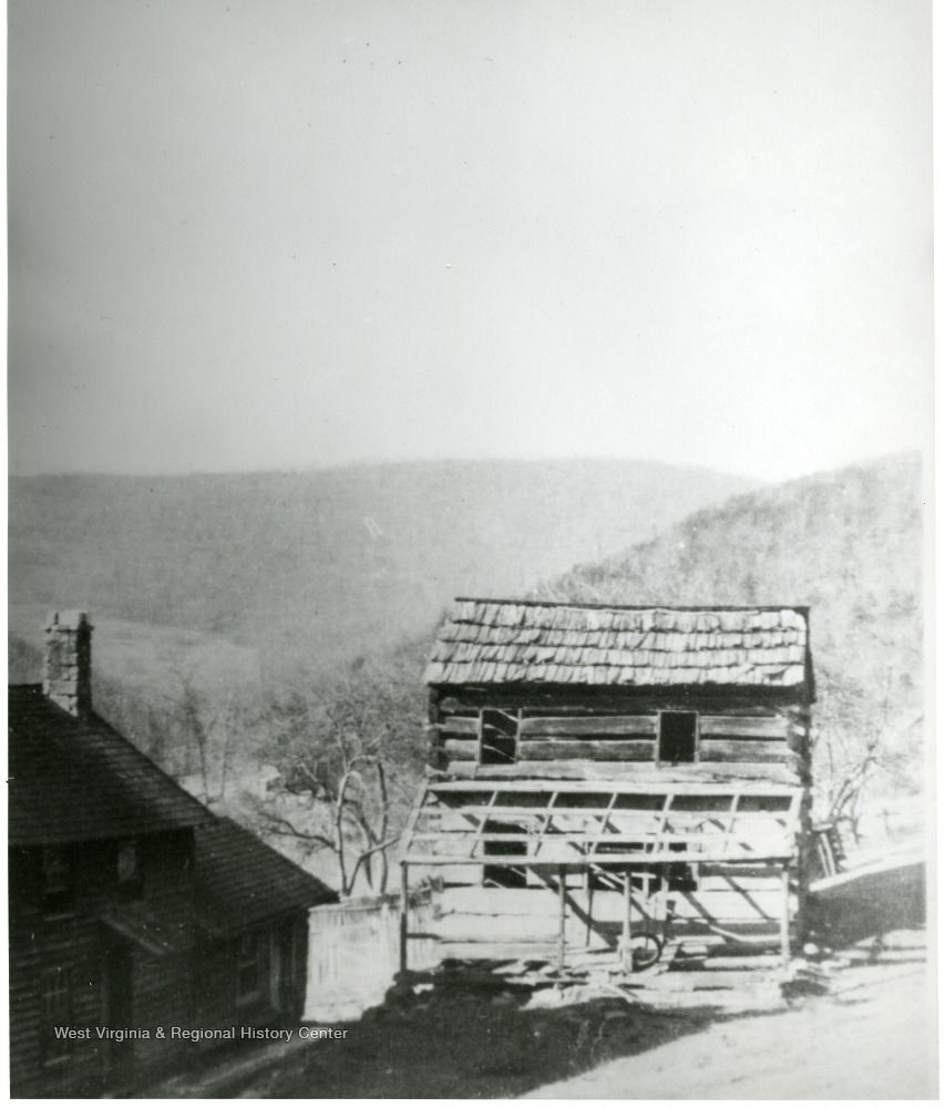 A vintage black-and-white photograph shows a rustic log cabin structure with an attached open porch against a hilly backdrop.