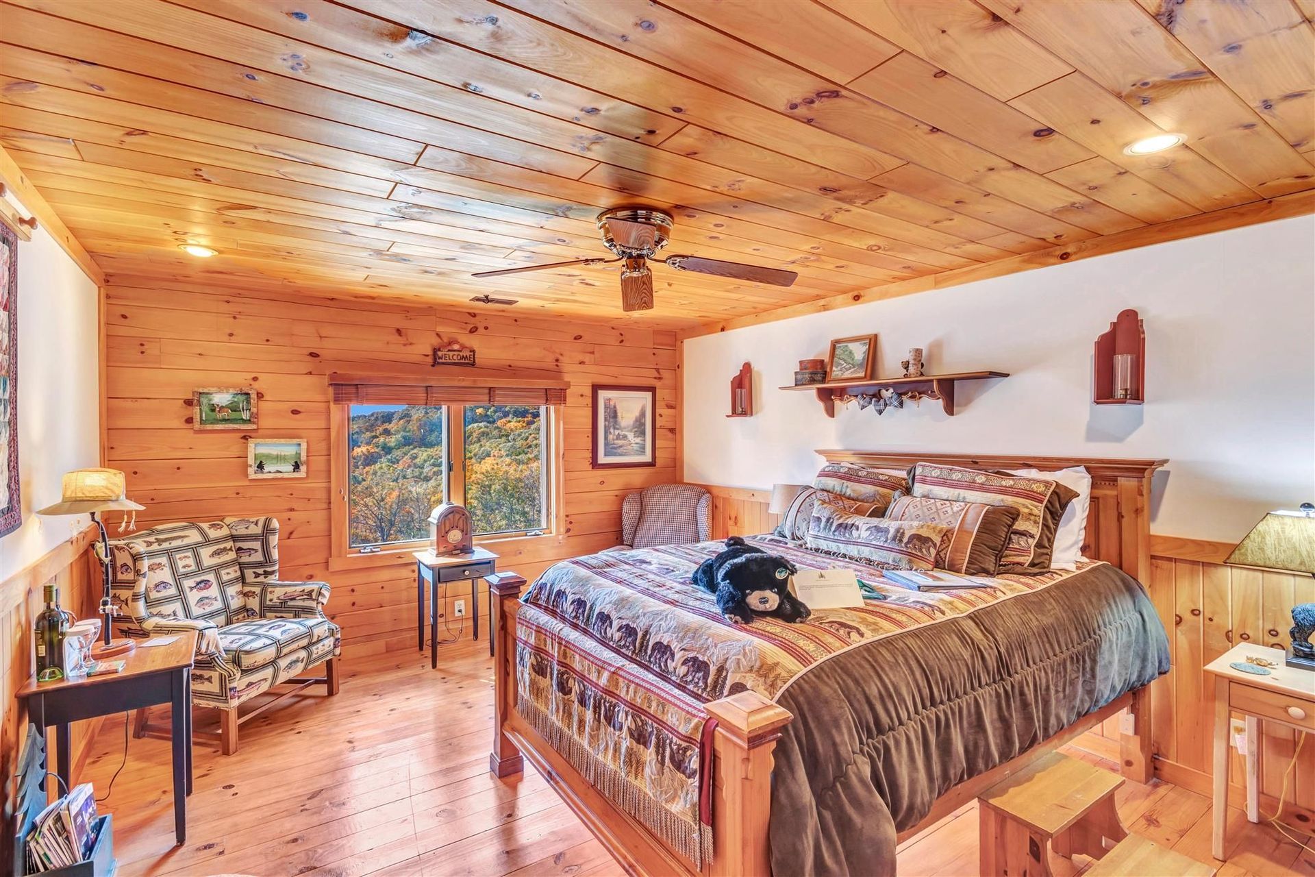 A rustic bedroom with wooden walls and ceiling, a large bed with a patterned quilt, a window, and an armchair.