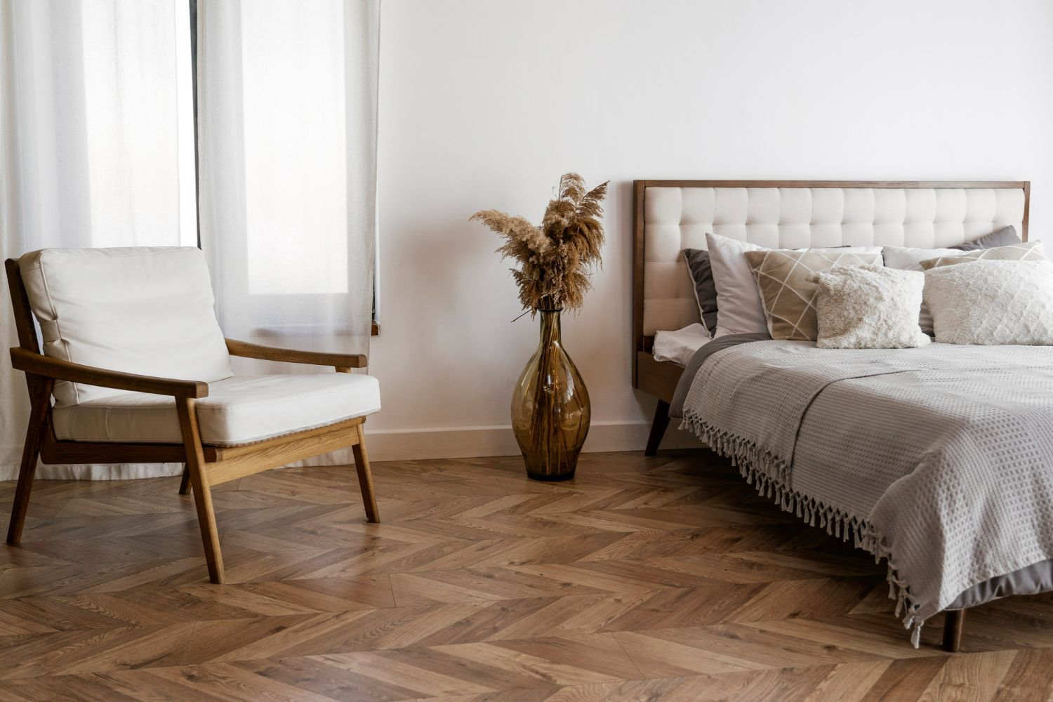 Bedroom interior with a bed, chair, vase of dried flowers, and wooden floor.