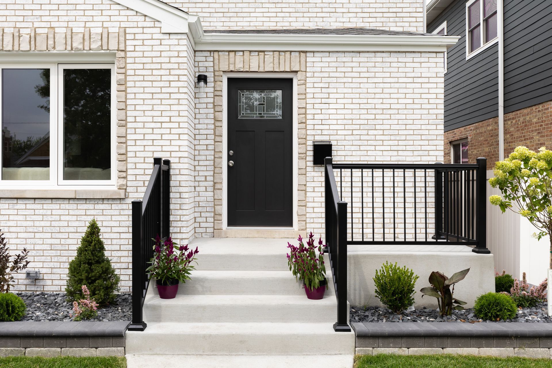 White brick house entrance with black door, steps, and railings. Landscaping includes bushes and flowers.