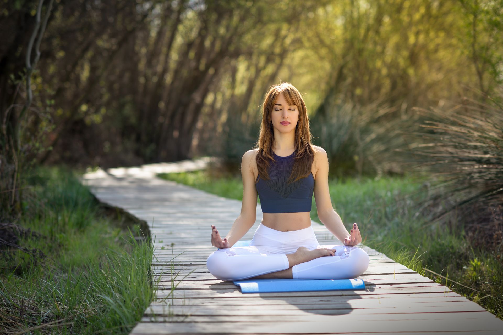 Woman meditates on a wooden path in a forest, eyes closed, hands in prayer pose