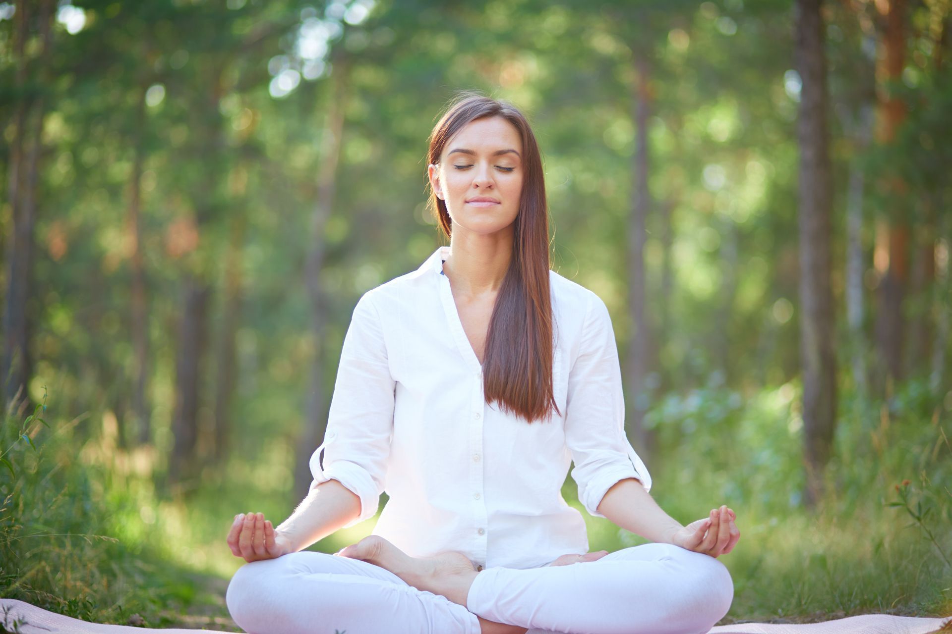 Woman meditating in a forest, eyes closed, hands in prayer pose, wearing white, surrounded by green trees