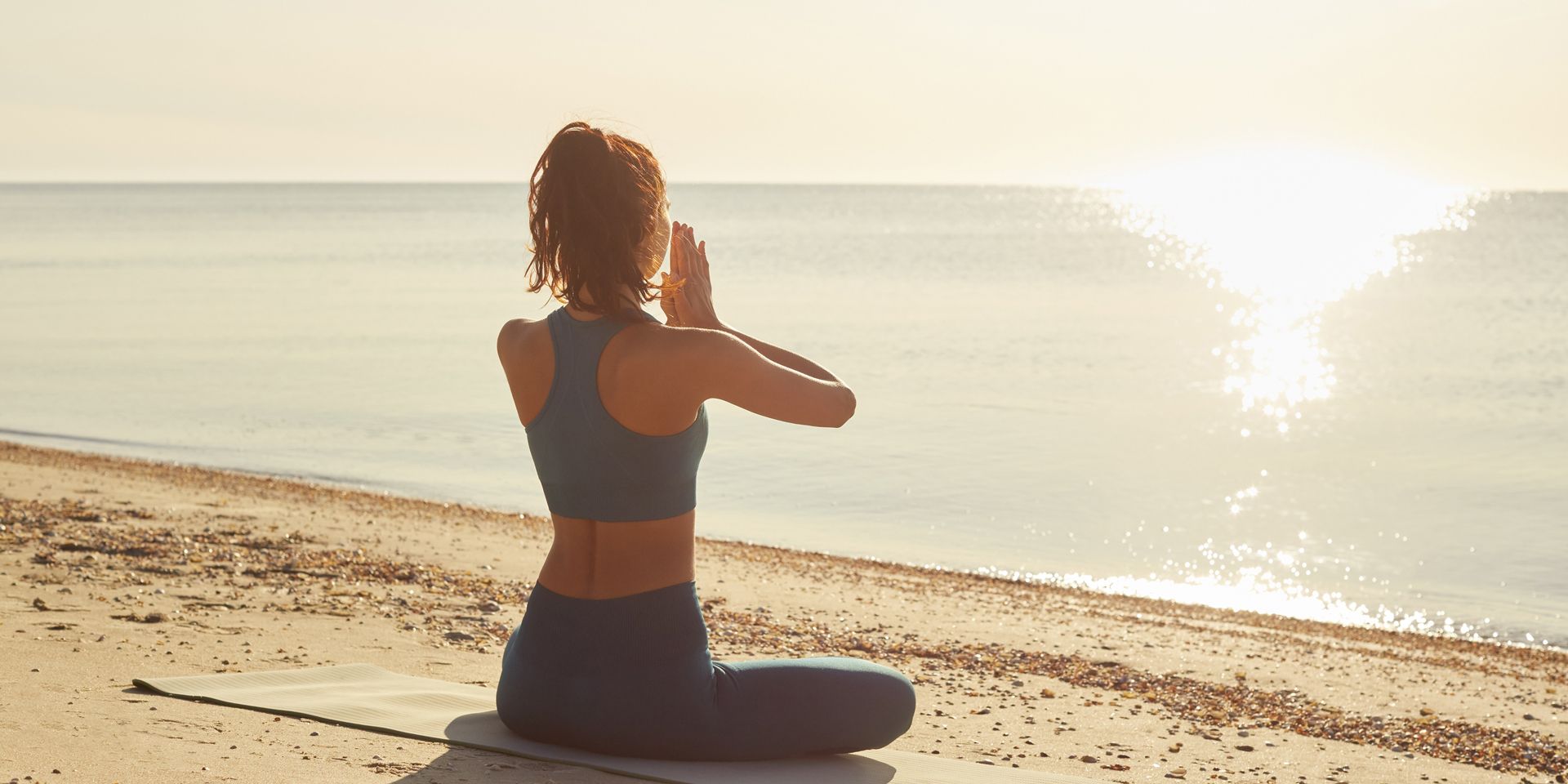 Woman in yoga pose on a beach at sunrise, hands together, back to viewer