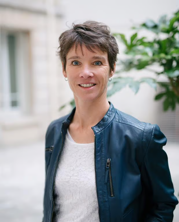 Une femme portant une veste en cuir bleu et une chemise blanche sourit à la caméra.