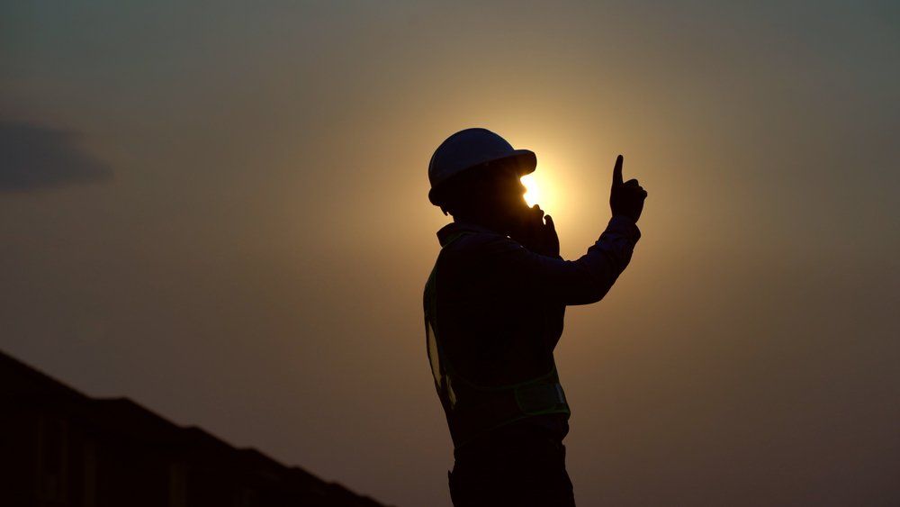 A silhouette of a man wearing a hard hat pointing at the sun at sunset.