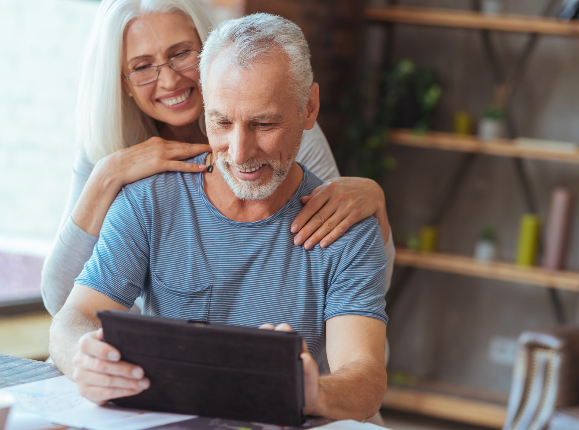An elderly couple is sitting at a table looking at a tablet.