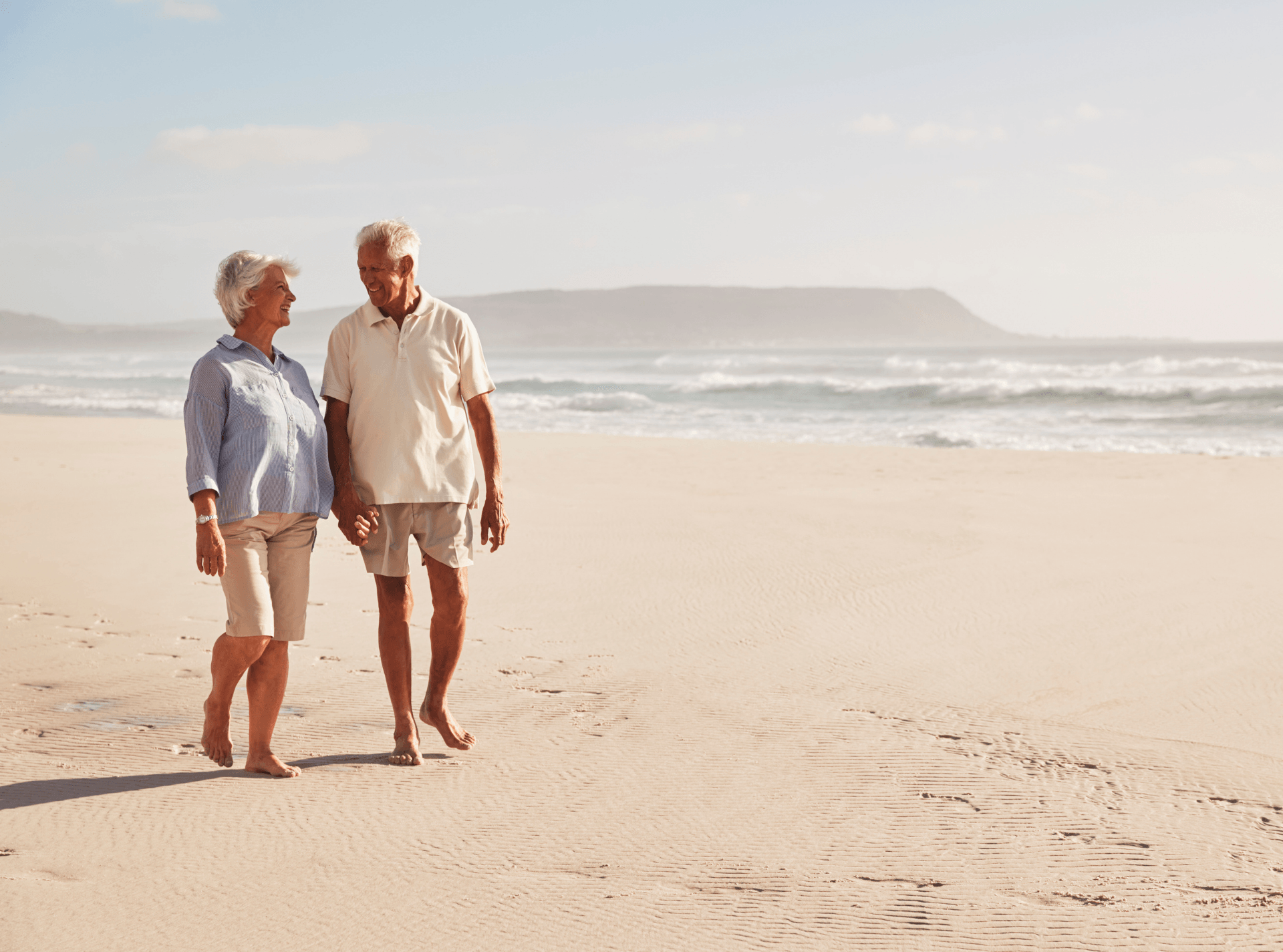 An elderly couple is walking on the beach holding hands.
