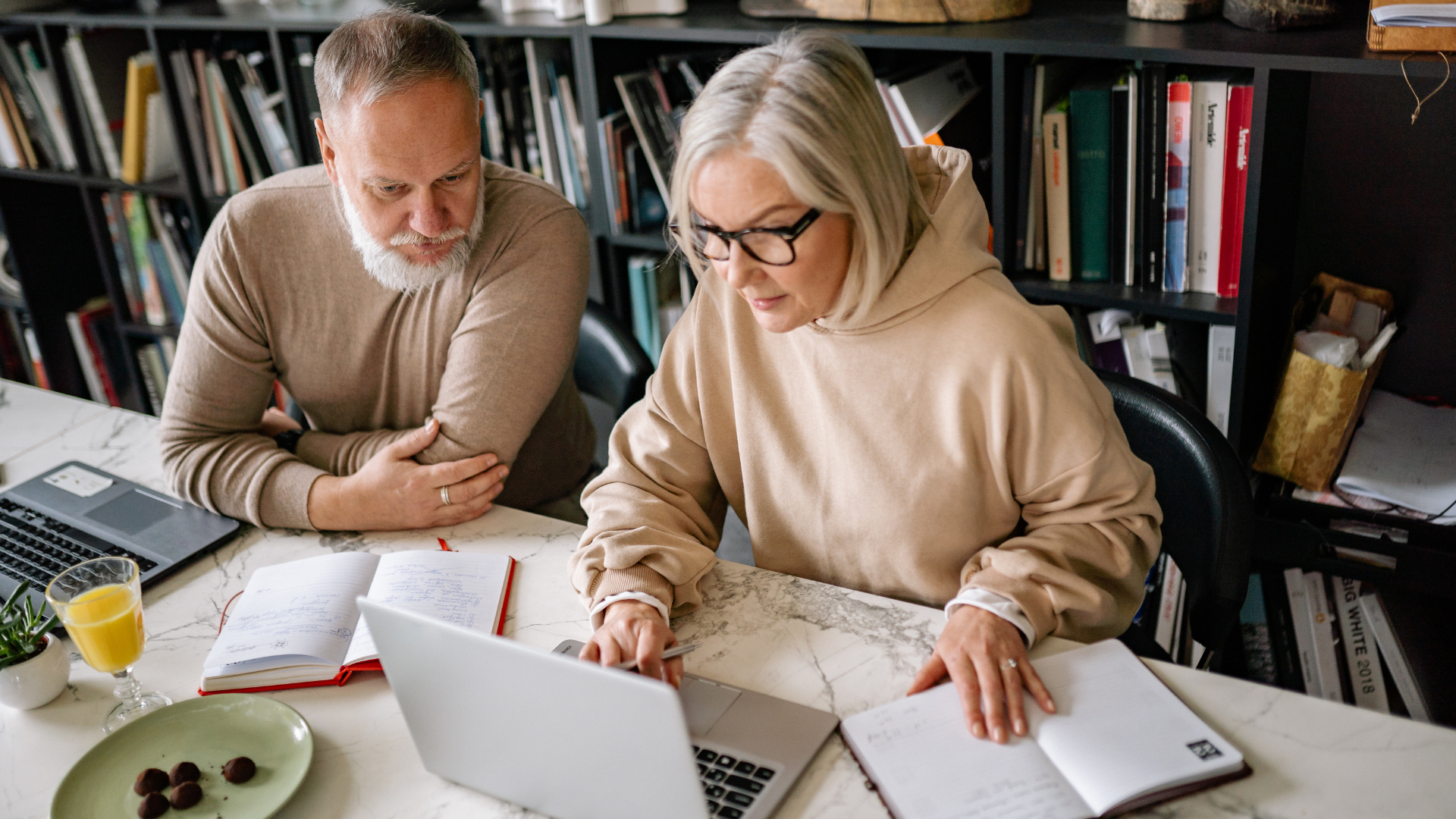 Two people sit at a table with books and a laptop, appearing to work or study together in a room with a large bookshelf.