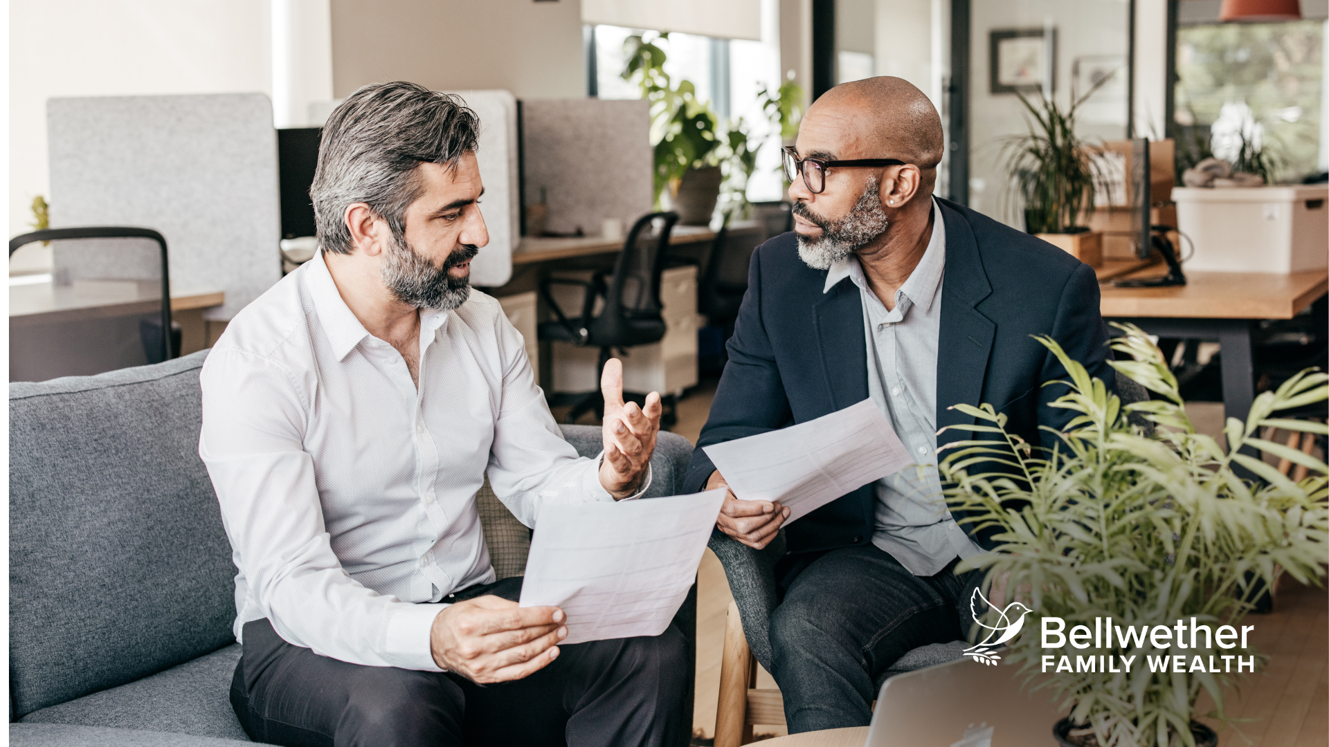 Two men are sitting on a couch talking to each other while holding papers.