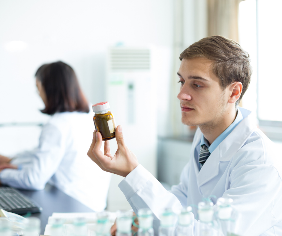 Man in lab coat examines a bottle in a lab setting. A person sits at a desk in the background.