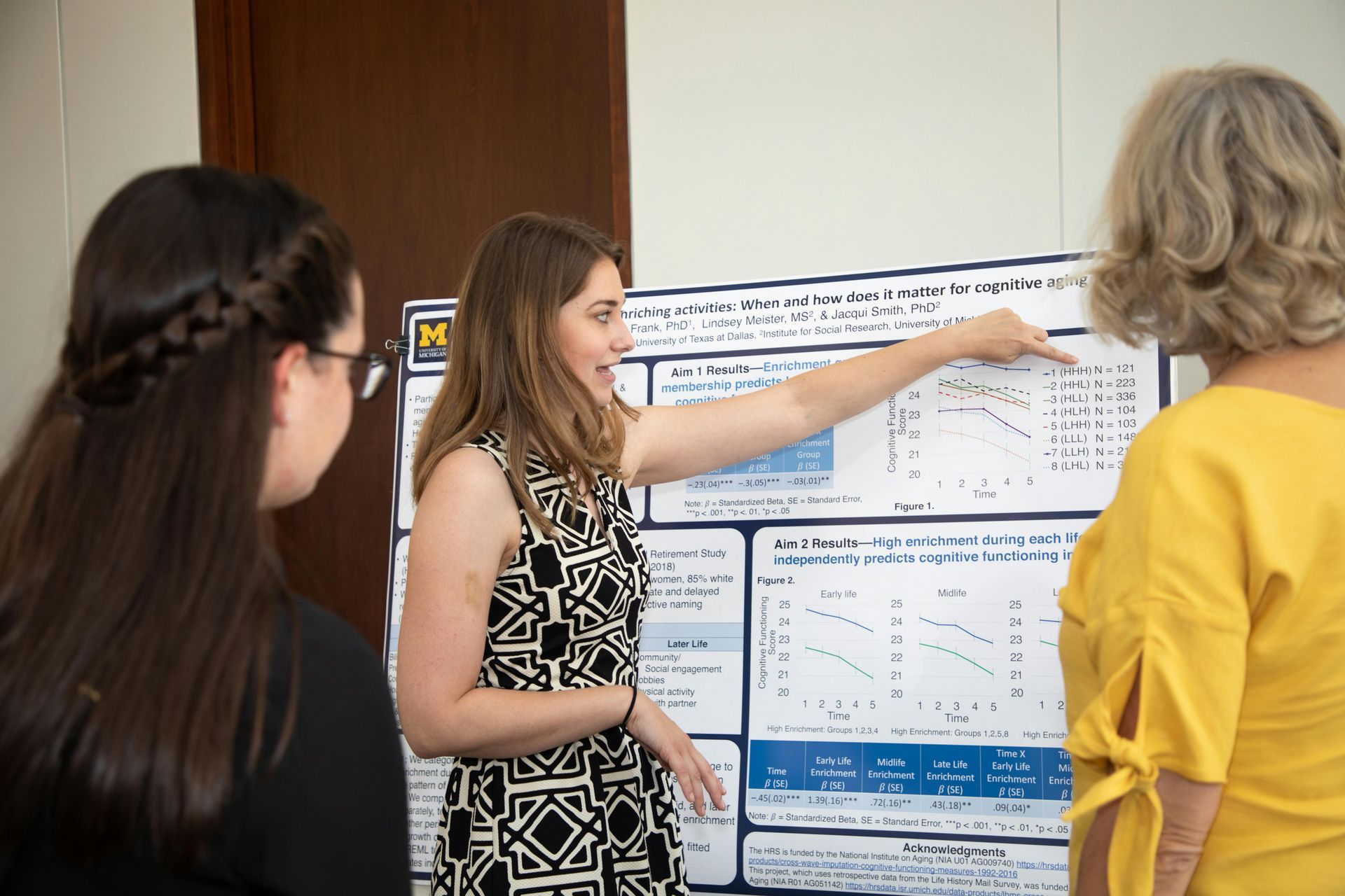 Woman presenting a research poster, pointing at a graph. Two others observe, one with braided hair.