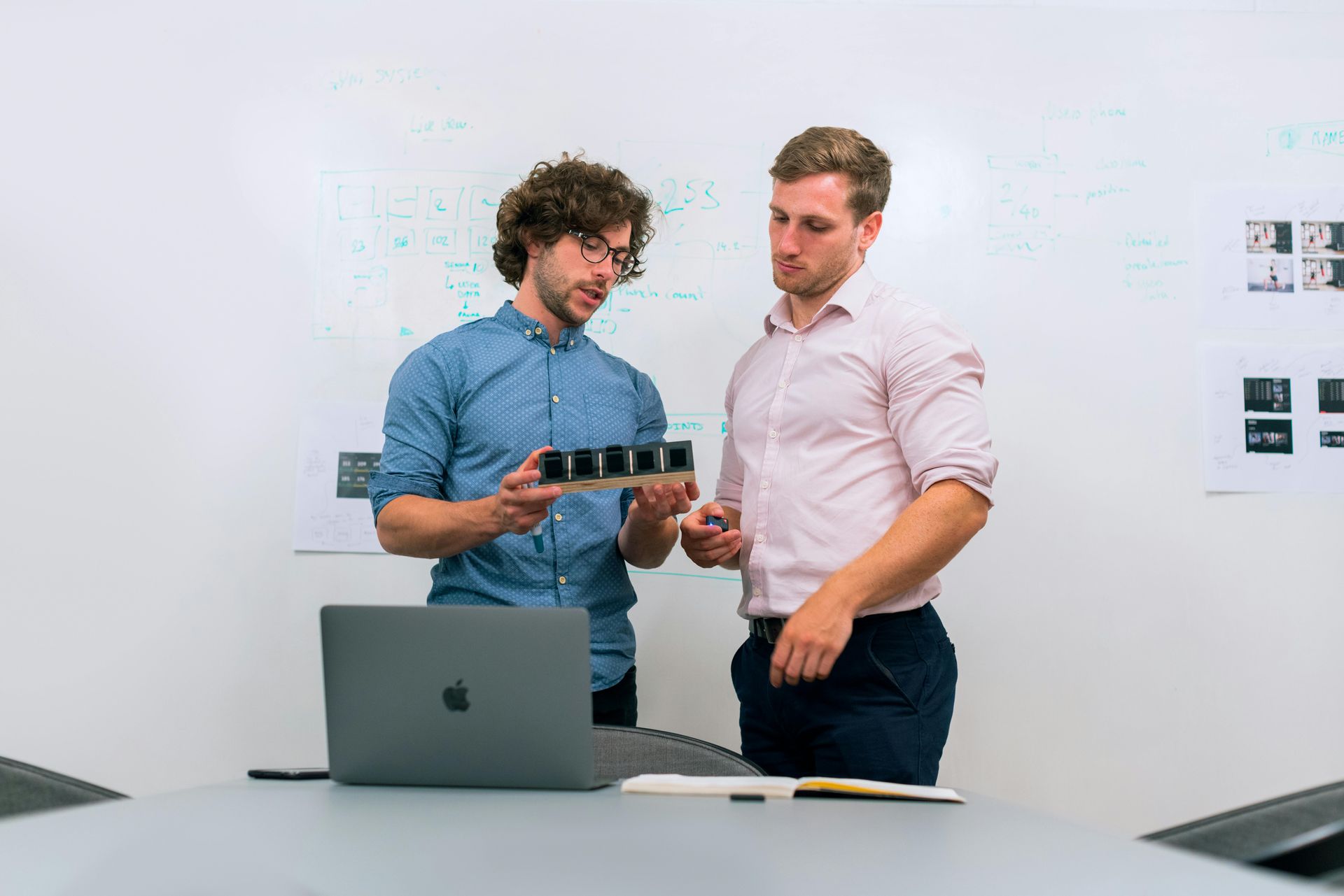 Two men examining a small, dark object; a laptop and whiteboard are in the background.
