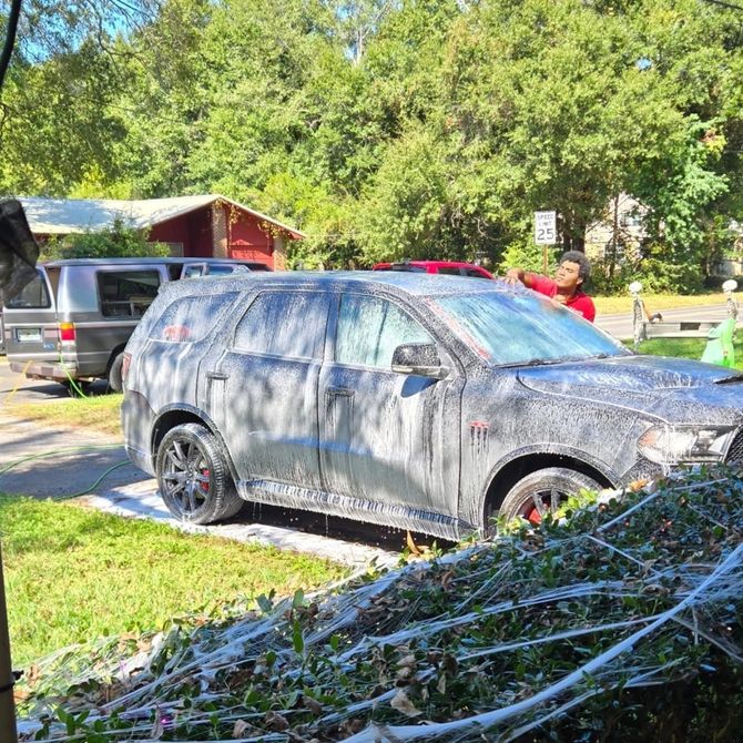 Person washing a black SUV with soap. Green lawn, trees, and other cars in the background.