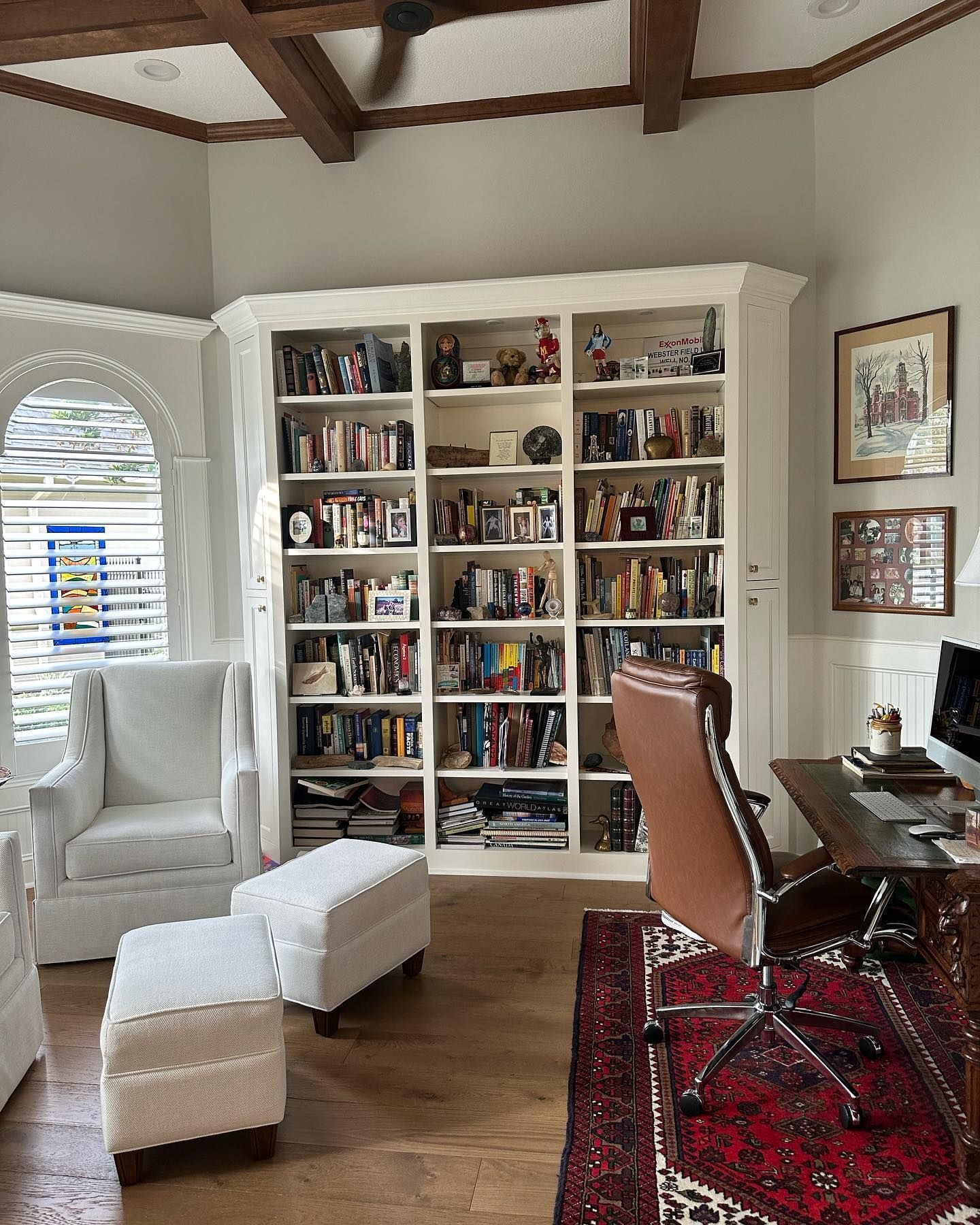 A living room with a large bookshelf filled with books , chairs , ottomans and a desk.