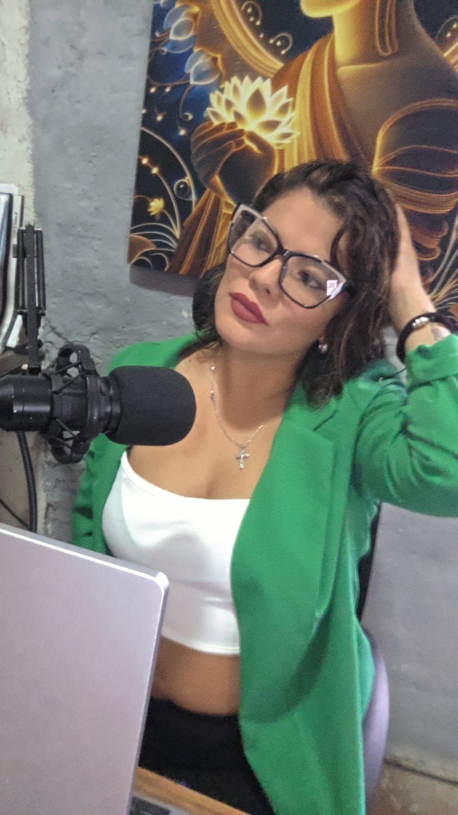 Una persona con gafas y chaqueta verde, sentada en un escritorio con un micrófono y un ordenador portátil, con la mano apoyada en la cabeza.