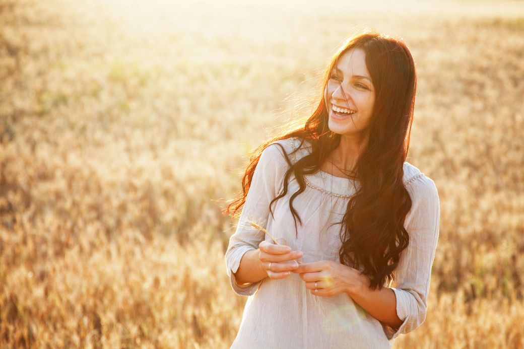 Una persona con cabello largo y oscuro, vestida con una blusa de color claro, sonríe mientras está de pie en un campo de trigo dorado al atardecer.