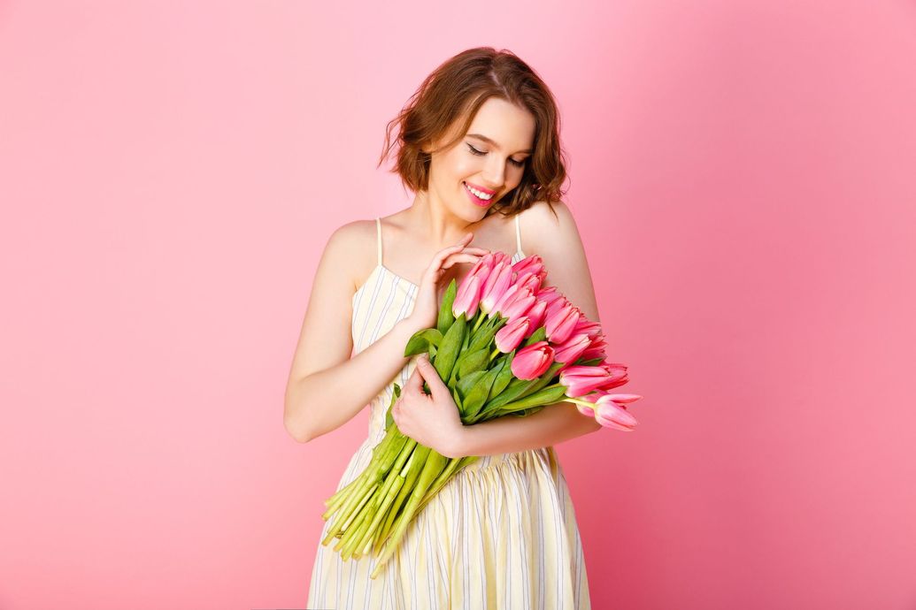 Una persona sonriente con un vestido color crema sostiene un ramo de tulipanes rosas sobre un fondo rosa liso.